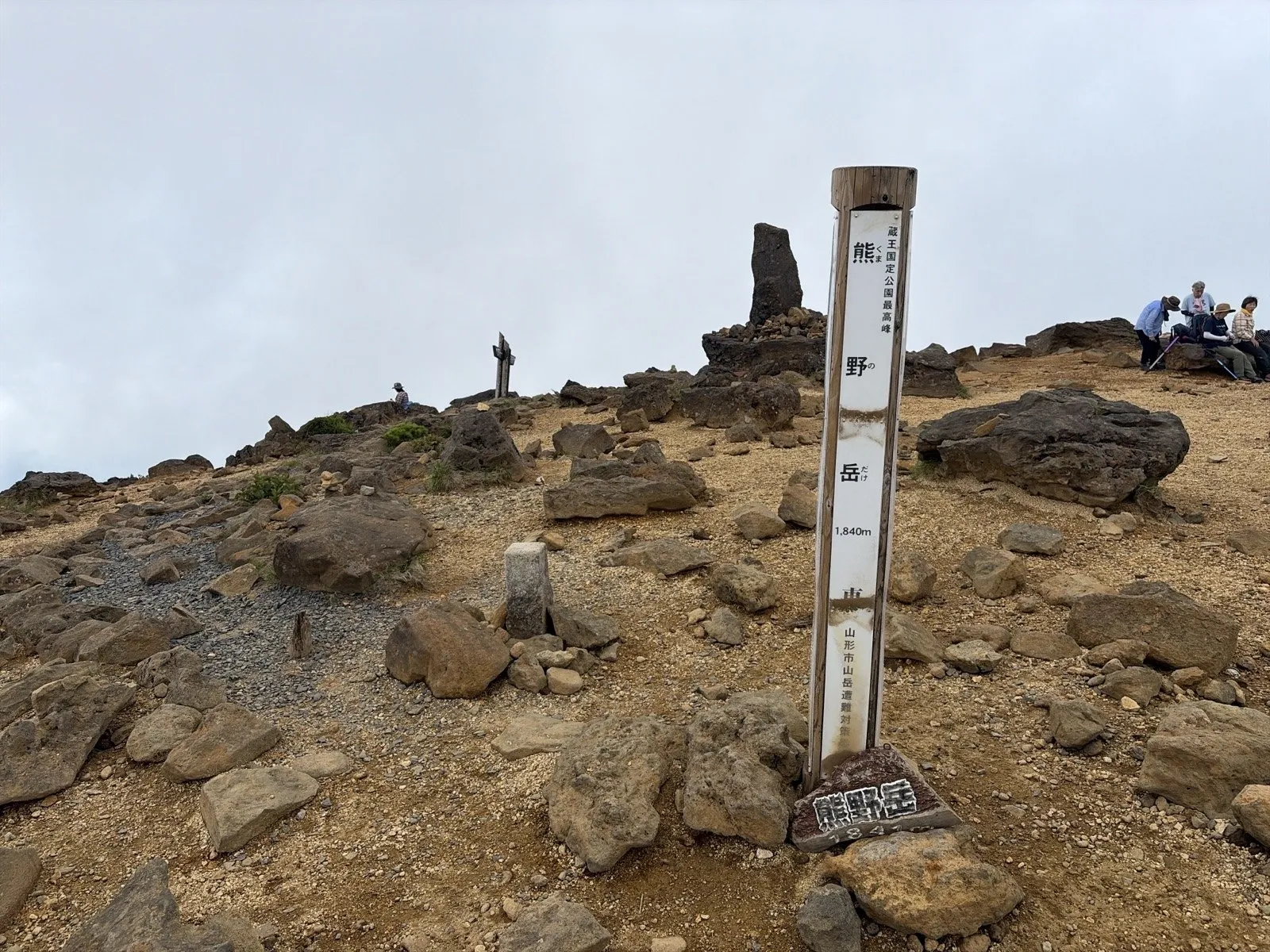 石が散らばる熊野岳の山頂に立つ白い標柱。奥に石積みと休憩する登山者が見え、まわりに木はない