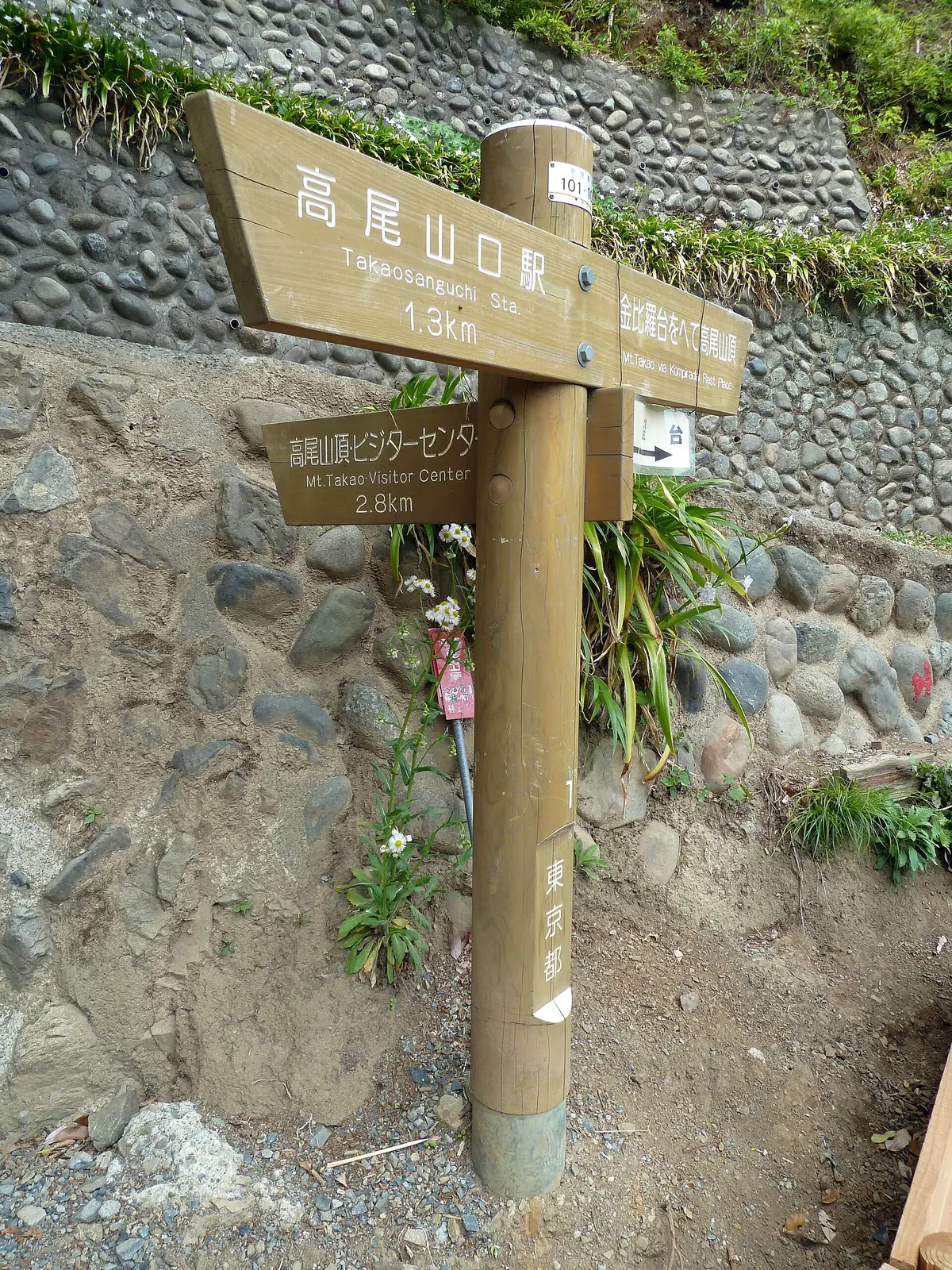 石垣の前に立つ木製の道標。高尾山口駅1.3km、高尾山ビジターセンター2.6kmと書かれている