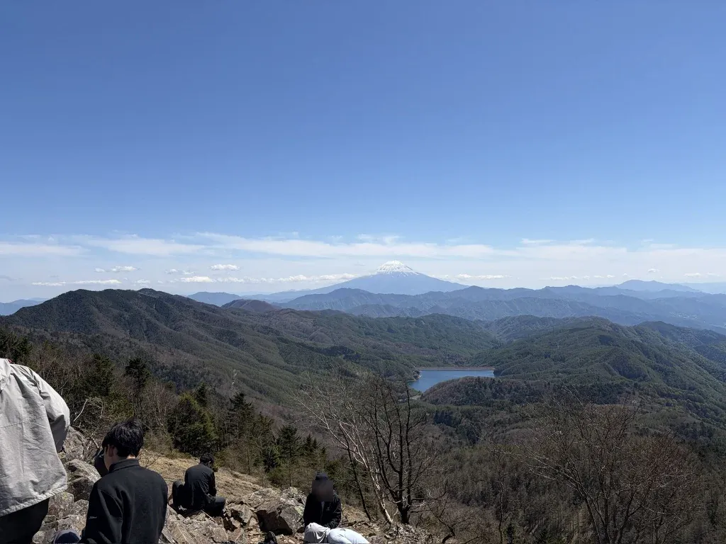 雷岩の岩の上で休憩する登山者と、正面に見える富士山と大菩薩湖