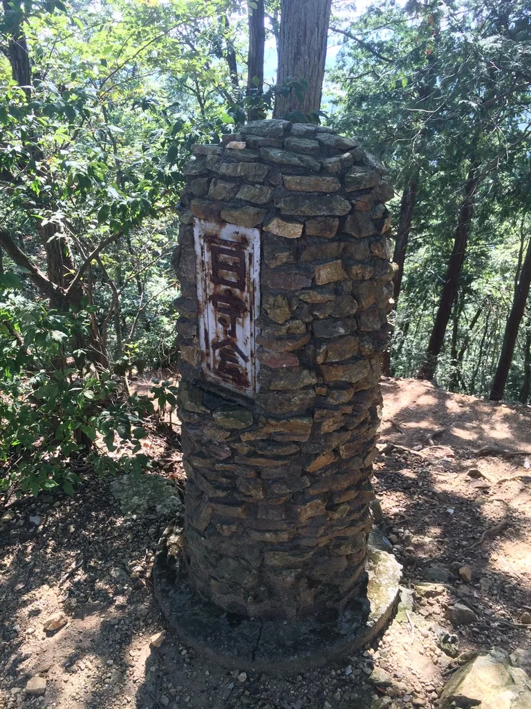 木々に囲まれた日和田山の山頂で、石を積み上げた塔と山名の板が立っている