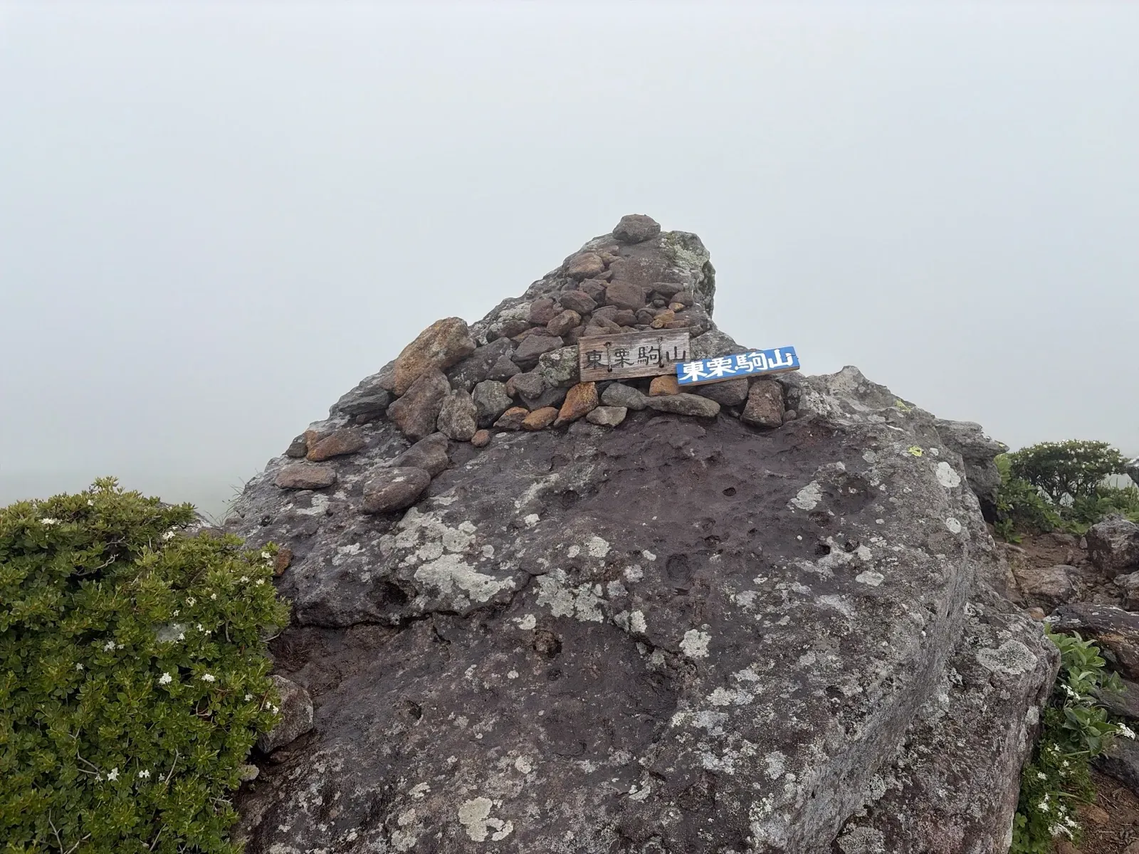 霧の中に立つ東栗駒山の山頂の岩。積まれた石に木と青色の「東栗駒山」の標識が付いている