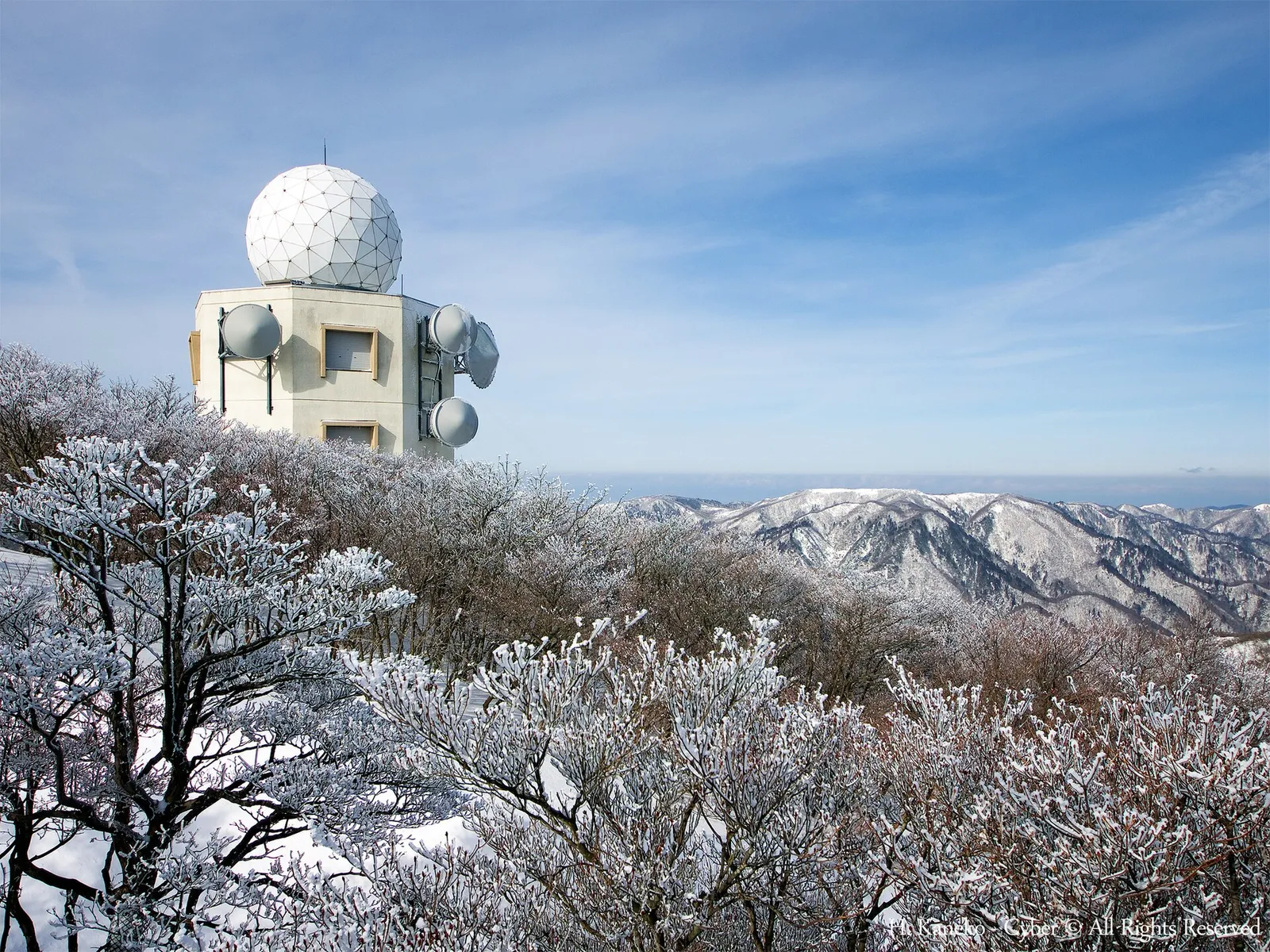 御在所岳の山頂付近で、白い霧氷に覆われた木々の向こうに白いドーム型の建物と、雪をかぶった鈴鹿山脈の山並みが見える