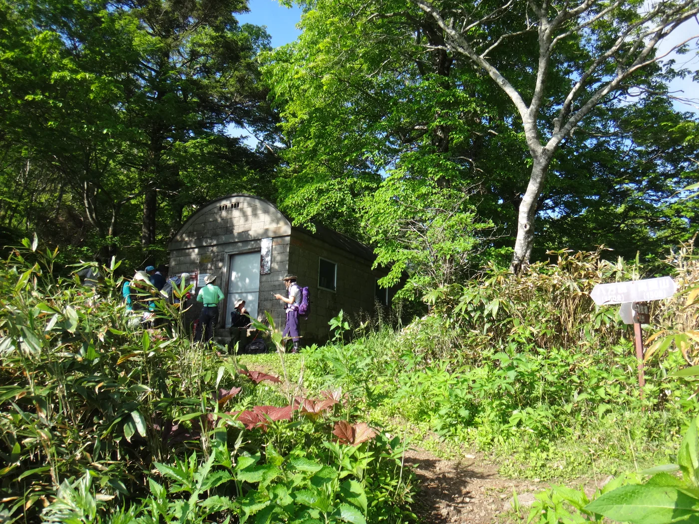樹林に囲まれた小さな石造りの一不動避難小屋と、その前で休む登山者