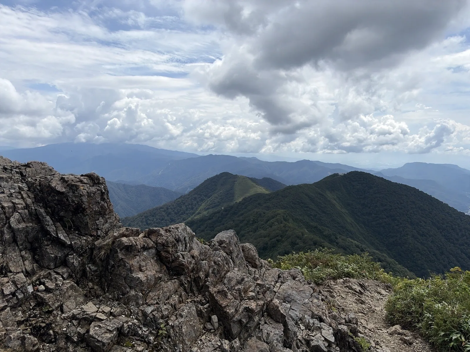 手前にごつごつした灰色の岩場があり、その向こうに緑の尾根と遠くの山並み、大きな雲が広がっている
