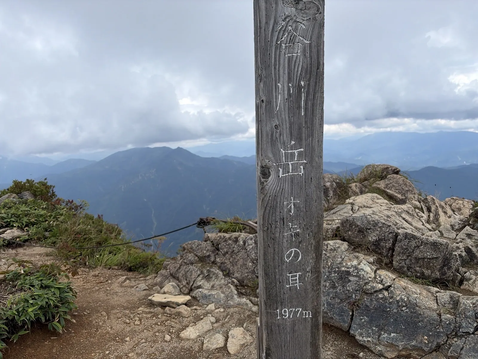 岩が積み重なるオキノ耳の山頂に立つ木の標柱。「谷川岳 オキの耳 1977m」と書かれ、奥には雲の下に山並みが広がっている