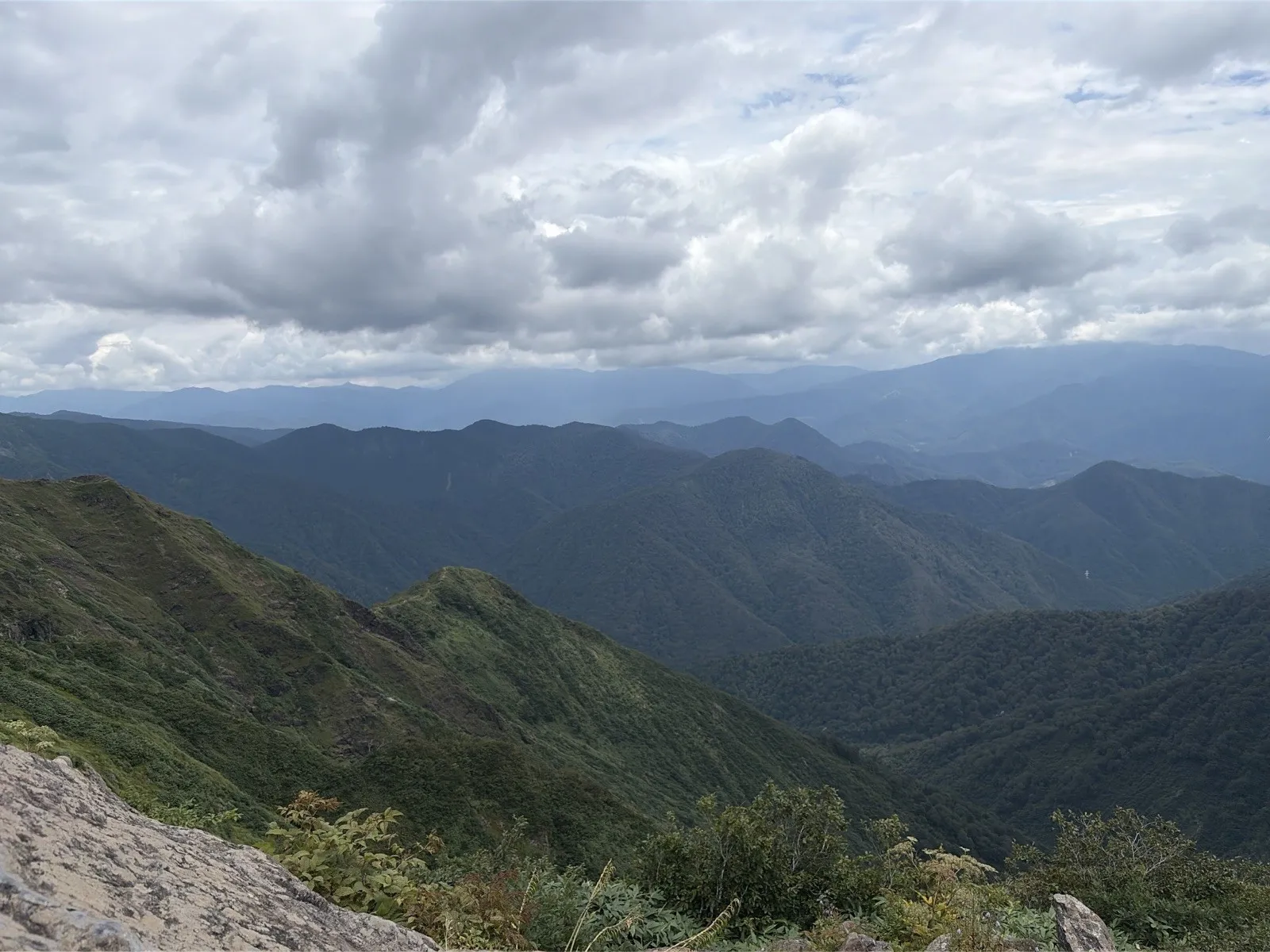 天狗の留まり場の岩の上から見下ろす、緑の尾根と幾重にも重なる山並み。空は厚い雲に覆われている