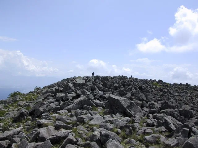 大きな溶岩塊が一面に広がる蓼科山の山頂部と、岩の上を歩く登山者